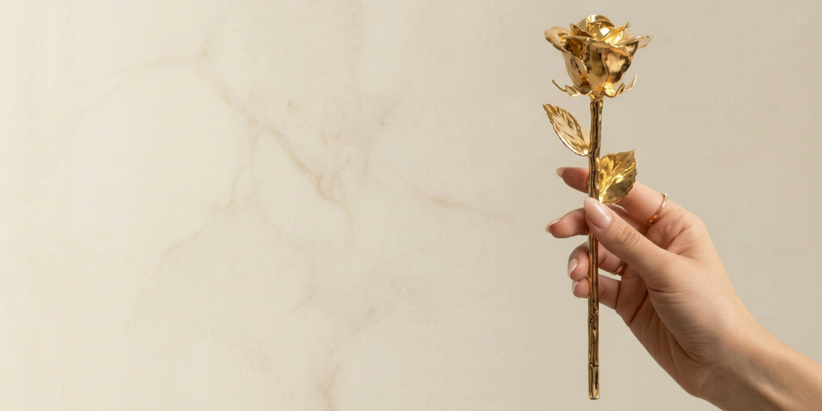 Hand holding a gold rose against a beige marble background